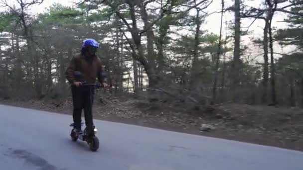 Kierowca noszący kask ochronny i potężny skuter elektryczny jeździ leśną drogą. Mężczyzna jeździ skuterem elektrycznym po lesie betonową drogą od strony kamery.. — Wideo stockowe