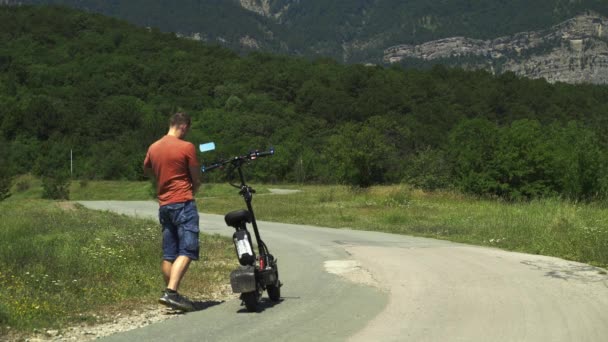 Rider ett kraftfullt elfordon. En man närmar sig en kraftfull elektrisk skoter, sätter på sig en skyddande hjälm och kör bort från kameran med ryggen mot bakgrund av en grön skog. — Stockvideo