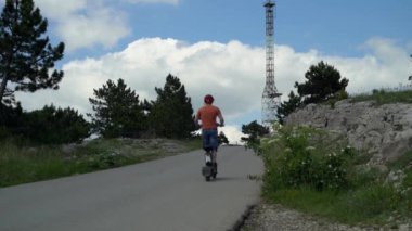 Güçlü bir elektrikli araca binmek. Elektrikli scooterlı koruyucu kasklı bir adam eski bir demir kulenin arka planına doğru yola çıkıyor.