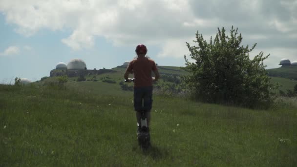 Op een krachtig elektrisch voertuig. Een man met een beschermende helm op een elektrische scooter rijdt op een groen veld tegen de achtergrond van een oud weerstation — Stockvideo