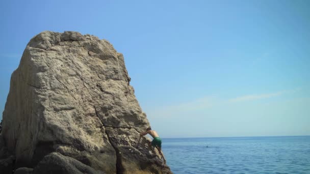 Junger Mann erklettert an einem heißen Sommertag einen hohen Felsen ins Meer, um ins Wasser zu springen — Stockvideo