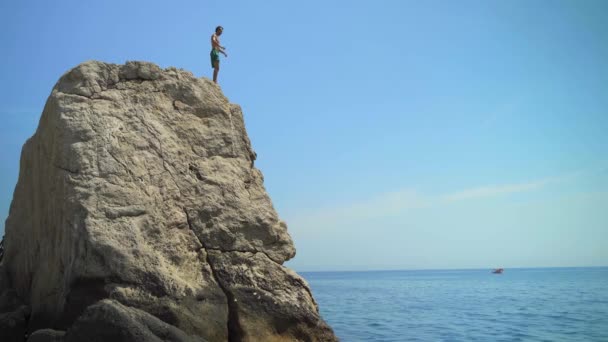 Ein junger Mann springt an einem heißen Sommertag von einer hohen Klippe ins Meer und macht einen Salto vorwärts. — Stockvideo