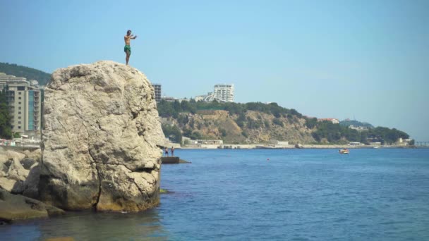 Um jovem salta de um penhasco alto para o mar fazendo belas curvas no ar e entra na água com os pés. — Vídeo de Stock