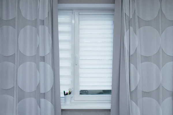 Odanın penceresinde beyaz panjurlar ve gri perdeler.
