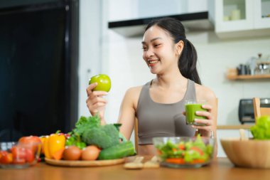 Asyalı kadın yeşil meyve suyu ve elma tutuyor, modern mutfakta taze sebze ve salata ile çevrili. Sağlıklı beslenme, zindelik ve sağlıklı yaşam tarzını teşvik eder..