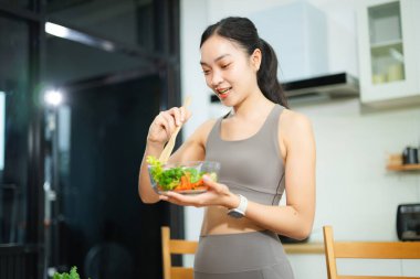 Gülümseyen, aktif giysili Asyalı kadın, mutfakta salata ve kırmızı elma tutuyor. Sağlıklı yaşam tarzı, zindelik, temiz yemek ve dengeli beslenmeyi teşvik eder..