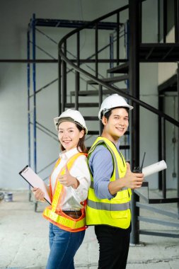 Tam teşekküllü proje planları ve dizüstü bilgisayarı üzerinde kutlama yapmak için iki sağlam şapka ve güvenlik yelekli inşaat profesyoneli yumruklarını kaldırarak sahada takım çalışması başarısını simgeliyor..