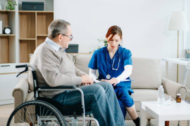 Mavi üniformalı profesyonel bir hemşire evde yaşlı bir tekerlekli sandalye hastasına danışmanlık yapıyor. Gerçek insanları, empatiyi ve kaliteli tıbbi tedaviyi sıcak ve destekleyici bir ortamda sergiliyor..