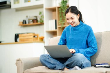 Genç Asyalı kadın rahat bir kanepede rahat rahat dizüstü bilgisayara mesaj atıyor ve tablet gülümseyen kız evden cep telefonu alışverişi yapıyor.