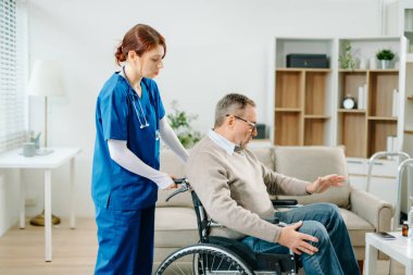 Mavi üniformalı profesyonel bir hemşire evde yaşlı bir tekerlekli sandalye hastasına danışmanlık yapıyor. Gerçek insanları, empatiyi ve kaliteli tıbbi tedaviyi sıcak ve destekleyici bir ortamda sergiliyor..