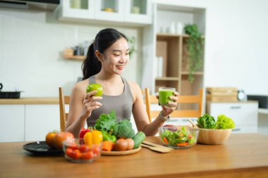 Asyalı kadın yeşil meyve suyu ve elma tutuyor, modern mutfakta taze sebze ve salata ile çevrili. Sağlıklı beslenme, zindelik ve sağlıklı yaşam tarzını teşvik eder..