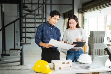Asyalı erkek ve kadın mimarlar kapalı bir inşaat alanının planlarını gözden geçiriyorlar. Modern bir mimari proje için takım çalışması, planlama ve mühendislik devam ediyor.