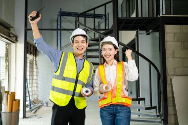 Tam teşekküllü proje planları ve dizüstü bilgisayarı üzerinde kutlama yapmak için iki sağlam şapka ve güvenlik yelekli inşaat profesyoneli yumruklarını kaldırarak sahada takım çalışması başarısını simgeliyor..