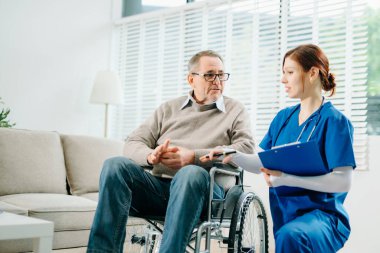 Mavi üniformalı profesyonel bir hemşire evde yaşlı bir tekerlekli sandalye hastasına danışmanlık yapıyor. Gerçek insanları, empatiyi ve kaliteli tıbbi tedaviyi sıcak ve destekleyici bir ortamda sergiliyor..