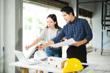 Asyalı erkek ve kadın mimarlar kapalı bir inşaat alanının planlarını gözden geçiriyorlar. Modern bir mimari proje için takım çalışması, planlama ve mühendislik devam ediyor.