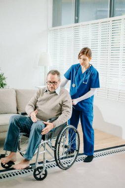 Mavi üniformalı profesyonel bir hemşire evde yaşlı bir tekerlekli sandalye hastasına danışmanlık yapıyor. Gerçek insanları, empatiyi ve kaliteli tıbbi tedaviyi sıcak ve destekleyici bir ortamda sergiliyor..