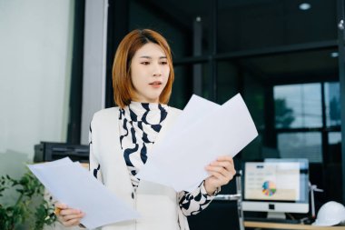 Genç Asyalı kadın ofiste kağıt raporlarını analiz ediyor, dizüstü bilgisayar ve belgelerle strateji planlıyor. İş, finans ve liderlik kavramları için ideal