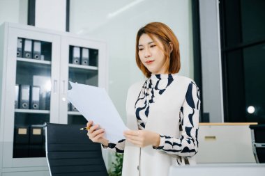 Genç Asyalı kadın ofiste kağıt raporlarını analiz ediyor, dizüstü bilgisayar ve belgelerle strateji planlıyor. İş, finans ve liderlik kavramları için ideal