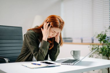 Genç Asyalı kadın dizüstü bilgisayarla çalışırken stres ve tükenme belirtileri gösteriyor. Aşırı çalışma, teslim tarihi baskısı ve iş yerinde akıl sağlığı kavramı.