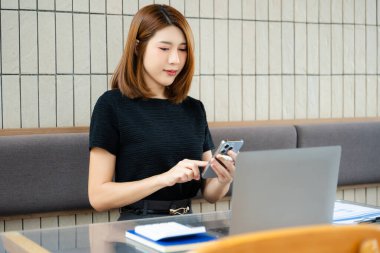 Asyalı iş kadını, modern bir ofiste hesap makinesiyle ve akıllı telefonuyla bir not panosundan mali durumu hesaplar., 