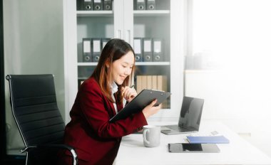 Çalışan not defteri, tablet ve dizüstü bilgisayarla çalışan Asyalı genç iş kadını akıllı telefon, tablet ve laptop video arama vergisi ile konuşuyor