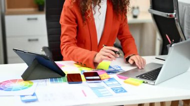 Ofisteki yaratıcı masalarda cihaz kullanan kadın grafik tasarımcılarına yakın çekim. Renk paletleri ve tasarım araçlarıyla çevrili 