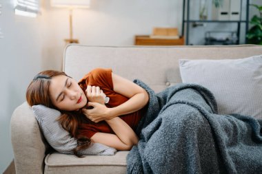 Kanepede battaniyeyle kaplanmış, grip belirtileri gösteren genç bir Asyalı kadın. Hastalık, sağlık ve iyileşme kavramı. Evde kal, sağlıklı kal.