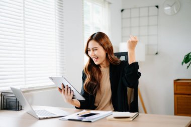 Asyalı iş kadını modern ofiste başarıyı kutluyor, tableti tutuyor ve dizüstü bilgisayarla gülümsüyor, iş, girişim ve dijital sektörde neşe ve başarı gösteriyor.