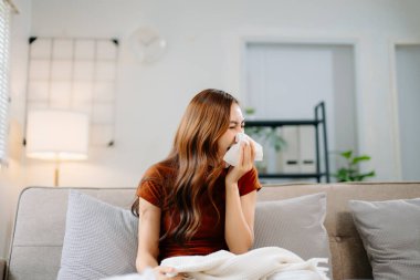 Kanepede battaniyeyle kaplanmış, grip belirtileri gösteren genç bir Asyalı kadın. Hastalık, sağlık ve iyileşme kavramı. Evde kal, sağlıklı kal.