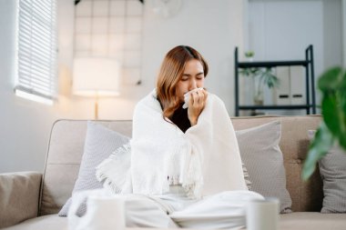 Kanepede battaniyeyle kaplanmış, grip belirtileri gösteren genç bir Asyalı kadın. Hastalık, sağlık ve iyileşme kavramı. Evde kal, sağlıklı kal.