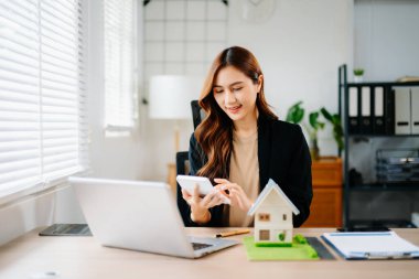 Ofisteki masada dizüstü bilgisayar ve tabletle çalışan genç bir emlakçı ve yanında da küçük bir ev..