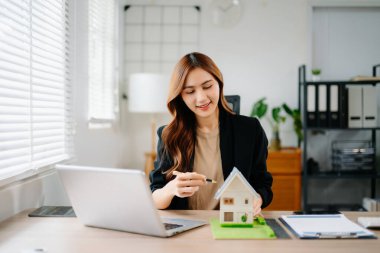Ofisteki masada dizüstü bilgisayar ve tabletle çalışan genç bir emlakçı ve yanında da küçük bir ev..