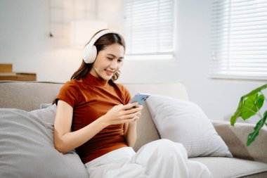 Kanepede oturan mutlu Asyalı kadın kulaklık takıyor ve akıllı telefon kullanıyor. Modern yaşam tarzı, sosyal medya ve çevrimiçi iletişim konsepti.