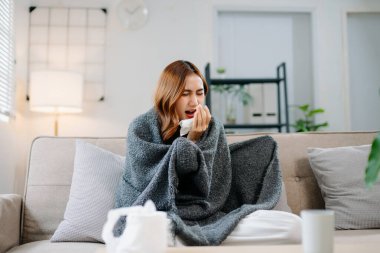 Kanepede battaniyeyle kaplanmış, grip belirtileri gösteren genç bir Asyalı kadın. Hastalık, sağlık ve iyileşme kavramı. Evde kal, sağlıklı kal.