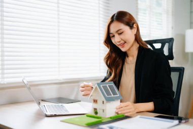 Ofisteki masada dizüstü bilgisayar ve tabletle çalışan genç bir emlakçı ve yanında da küçük bir ev..