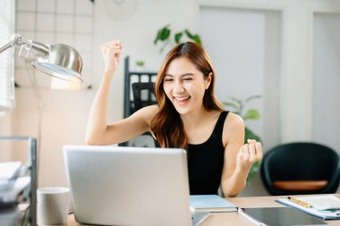 Neşeli iş kadını işyerinde başarıyı kutluyor, dizüstü bilgisayarda sevinç yumruklarını kaldırıyor, başarıyı, motivasyonu ve kariyer mutluluğunu simgeliyor..