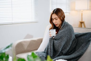 Kanepede battaniyeyle kaplanmış, grip belirtileri gösteren genç bir Asyalı kadın. Hastalık, sağlık ve iyileşme kavramı. Evde kal, sağlıklı kal.