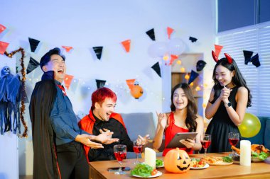 Cadılar Bayramı 'nda balkabakları, yiyecek ve içeceklerle dolu bir masada gülen ve yemek yiyen cadı ve şeytan kostümlü bir grup arkadaş..