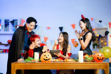 Cadılar Bayramı 'nda balkabakları, yiyecek ve içeceklerle dolu bir masada gülen ve yemek yiyen cadı ve şeytan kostümlü bir grup arkadaş..