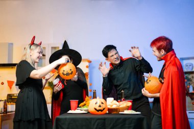 Cadılar Bayramı kostümlü bir grup arkadaş şenlikli bir partide ürkütücü aksesuar, fener ve kafatası dekoruyla gülüp oynuyorlar. Gerçek insanlar, yemek, içki ve neşe.