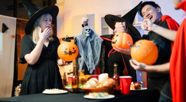 Cadılar Bayramı kostümlü bir grup arkadaş şenlikli bir partide ürkütücü aksesuar, fener ve kafatası dekoruyla gülüp oynuyorlar. Gerçek insanlar, yemek, içki ve neşe.