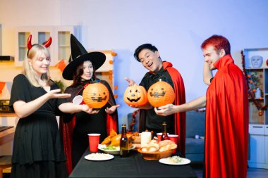 Cadılar Bayramı kostümlü bir grup arkadaş şenlikli bir partide ürkütücü aksesuar, fener ve kafatası dekoruyla gülüp oynuyorlar. Gerçek insanlar, yemek, içki ve neşe.