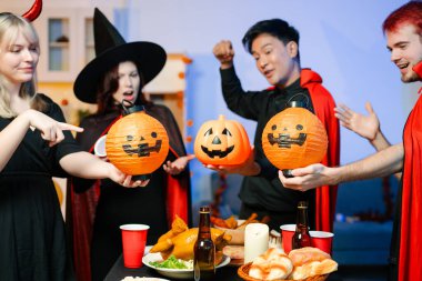 Cadılar Bayramı kostümlü bir grup arkadaş şenlikli bir partide ürkütücü aksesuar, fener ve kafatası dekoruyla gülüp oynuyorlar. Gerçek insanlar, yemek, içki ve neşe.