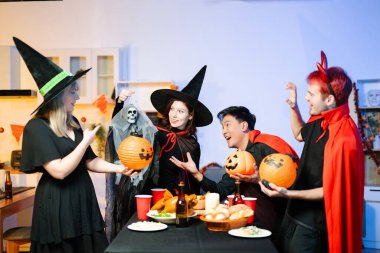 Cadılar Bayramı kostümlü bir grup arkadaş şenlikli bir partide ürkütücü aksesuar, fener ve kafatası dekoruyla gülüp oynuyorlar. Gerçek insanlar, yemek, içki ve neşe.