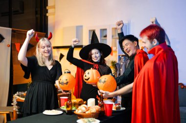 Cadılar Bayramı kostümlü bir grup arkadaş şenlikli bir partide ürkütücü aksesuar, fener ve kafatası dekoruyla gülüp oynuyorlar. Gerçek insanlar, yemek, içki ve neşe.