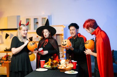 Cadılar Bayramı kostümlü bir grup arkadaş şenlikli bir partide ürkütücü aksesuar, fener ve kafatası dekoruyla gülüp oynuyorlar. Gerçek insanlar, yemek, içki ve neşe.