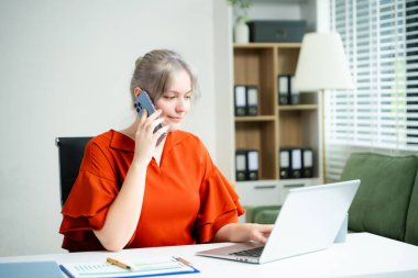 Uzaktan çalışan genç profesyonel bir kadın. Aydınlık bir ofiste dizüstü bilgisayar ve telefonla birden fazla iş yapıyor. İletişim, serbest çalışma ve modern iş hayatı kavramı.