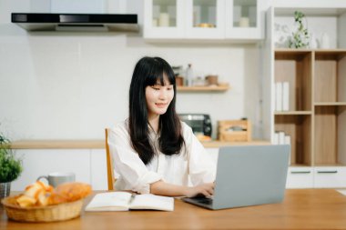 Asyalı kadın rahat bir mutfak çalışma alanında dizüstü bilgisayar ve tablet üzerinde çalışıyor. Eğitim, serbest meslek ve yenilik temaları için mükemmel