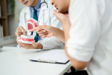 Diş hekimi, erkek hastaya diş sağlığını model çene kullanarak açıklıyor. Modern klinikte ağız bakımı, diş ağrısı danışmanlığı, anatomi eğitimi ve hijyen tavsiyesi.