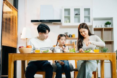 Asyalı aile kahvaltı saatini paylaşıyor. Ebeveynler, sıcak bir mutfakta yemek ve akıllı telefondan birbirlerine bağlanırken kızının süt içmesine yardım ediyorlar. Sıcak, samimi ebeveynlik anı.